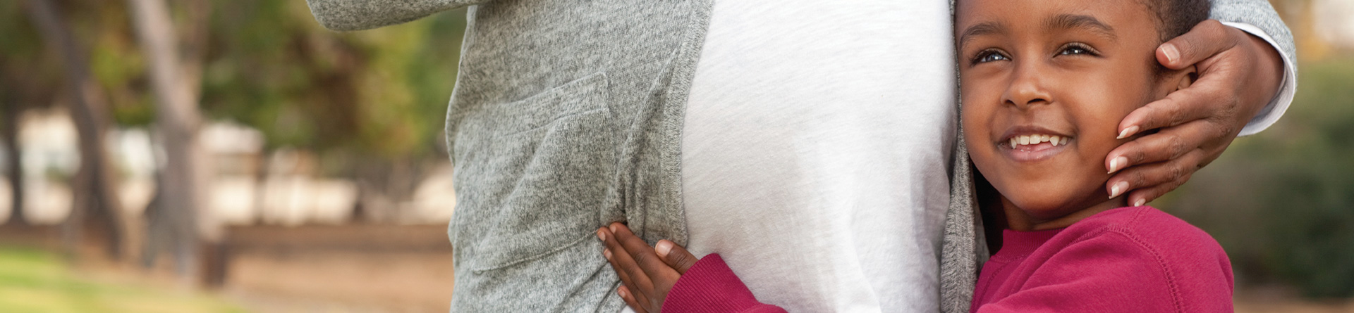 Child hugging pregnant parent outdoors, smiling warmly